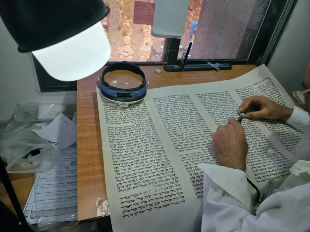 סופר סת"ם בודק יריעת קלף של ספר תורה תחת זכוכית מגדלת ומנורה