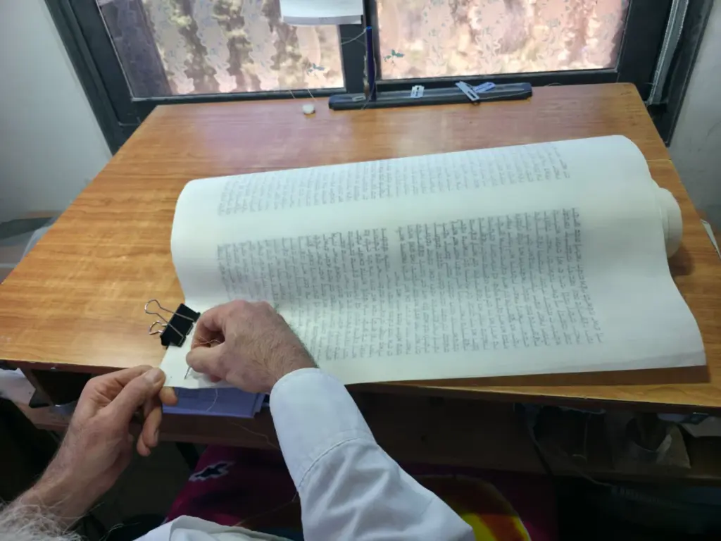 יריעת קלף של ספר תורה פרושה על שולחן עבודה ליד חלון